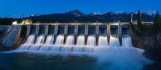 a large dam with water pouring over it and mountains in the backgroung