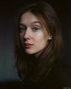 a woman with blue eyes and long brown hair is looking at the camera while standing in front of a dark background