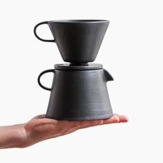 a hand holding a black coffee pot on top of a white background with one cup in the middle