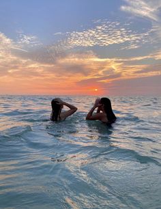 two women in the ocean at sunset with their backs to each other, one laying on her head