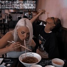 two women sitting at a table with chopsticks in their mouths and eating noodles