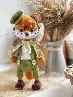 a crocheted stuffed animal wearing glasses and a green outfit next to a vase with dried flowers