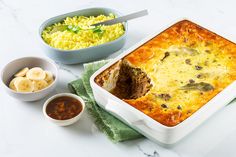 a casserole dish with meat, cheese and vegetables next to bowls of rice