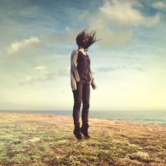 a woman standing on top of a grass covered field next to the ocean with her hair blowing in the wind