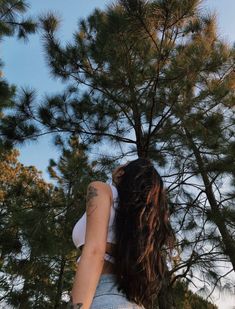 a woman with long hair standing in front of pine trees looking up at the sky