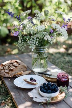 a table topped with blueberries and bread next to a vase filled with flowers on top of a wooden board