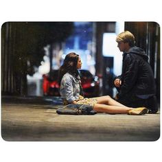 a young man and woman sitting on the ground next to each other looking at each other