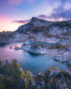 a mountain lake surrounded by trees and rocks at sunset with the sun setting in the distance