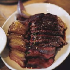 a white plate topped with sliced up meat next to a bowl filled with sauce and onions