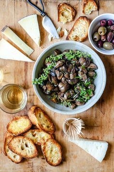 a bowl filled with mushrooms next to bread and olives on top of a wooden table