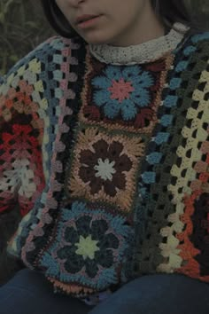 a woman wearing a multicolored crocheted sweater sitting in the grass with her hands on her hips