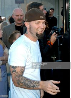 a man in white shirt and black hat smiling at the camera with people behind him