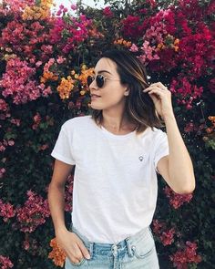 a woman in white shirt and jeans standing next to flower bush with sunglasses on her head