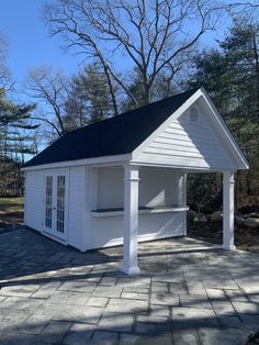 a small white building sitting on top of a brick patio next to a tree filled forest