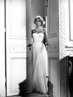 an old photo of a woman in a white dress standing by a door and looking at the camera