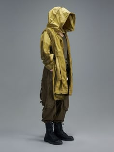 a man in a yellow raincoat and black boots stands against a gray background with his back turned to the camera