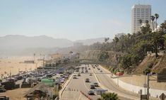 cars are driving on the highway near the beach and mountains in the distance, with palm trees lining the road