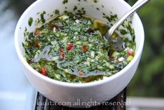 a white bowl filled with lots of green food next to a metal spoon on top of a table