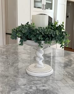 a white vase with green plants in it sitting on a marble counter top next to a candle