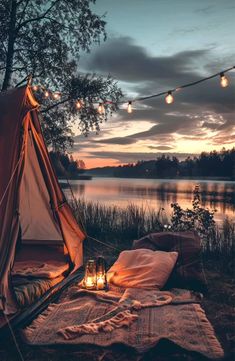 a tent is set up next to a lake with lights strung over it and blankets on the ground