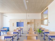 an empty classroom with wooden desks and blue chairs in front of a wall mounted television