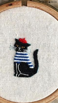 a black cat with a red hat and striped shirt is sitting in front of an embroidery hoop