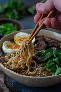 a person holding chopsticks over a bowl of noodles with meat and egg in it