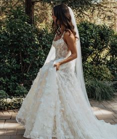 a woman wearing a wedding dress and veil walking down the street in front of bushes