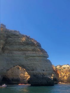 two boats are in the water near large rocks and cliffs on a clear blue sky day