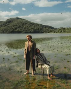 two men standing next to each other in front of a body of water with mountains in the background