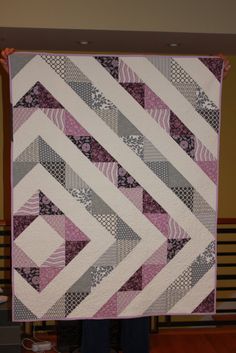 a person holding up a quilt made with squares and strips in pink, grey and white