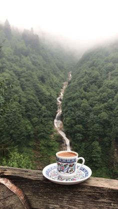 a cup sitting on top of a wooden table next to a lush green forest covered hillside