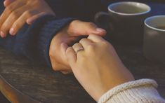 two people holding hands over a table with coffee cups in front of them and one person's hand on the table