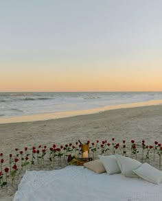 a white blanket on the beach with red roses and candles in front of it at sunset