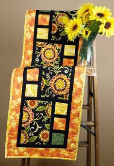 a vase filled with sunflowers sitting on top of a wooden chair next to a black and yellow quilt