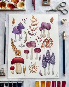 watercolors and paint supplies are laid out on a table with mushrooms, leaves, and flowers