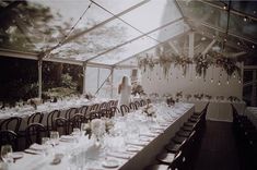 a long table is set up for a wedding reception in a glass walled room with white linens and greenery hanging from the ceiling