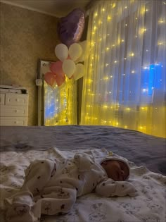 a baby laying on top of a bed next to a window with balloons hanging from it