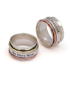 two silver and gold rings with the words be here now written on them, sitting next to each other