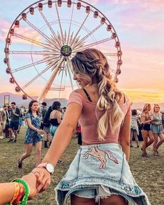 two girls holding hands in front of a ferris wheel at an outdoor music festival with other people