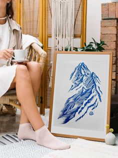 a woman sitting in a chair holding a cup and looking at a painting on the floor