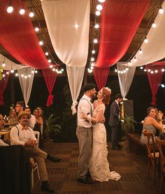 a bride and groom kissing in front of their guests at a wedding reception with red drapes hanging from the ceiling