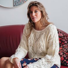 a woman sitting on top of a red couch wearing a white sweater and floral skirt