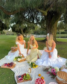 three women sitting on a blanket in front of a tree with food and drinks around them