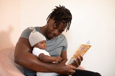 a man sitting in a chair holding a baby while reading a book