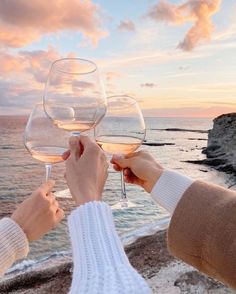 two people toasting wine glasses with the ocean and sky in the background at sunset