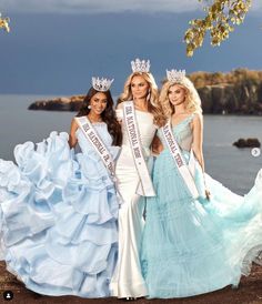 three beautiful women standing next to each other in dresses and tiaras on the beach