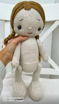 a hand holding a white knitted doll in front of a white chair with a brown hair