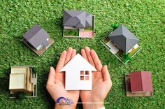 two hands are holding a paper house in front of several model houses on the grass