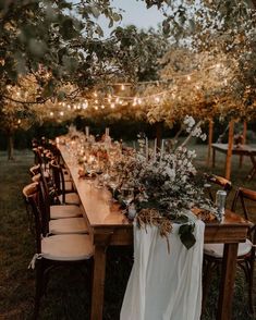 a long wooden table topped with lots of flowers and greenery next to string lights